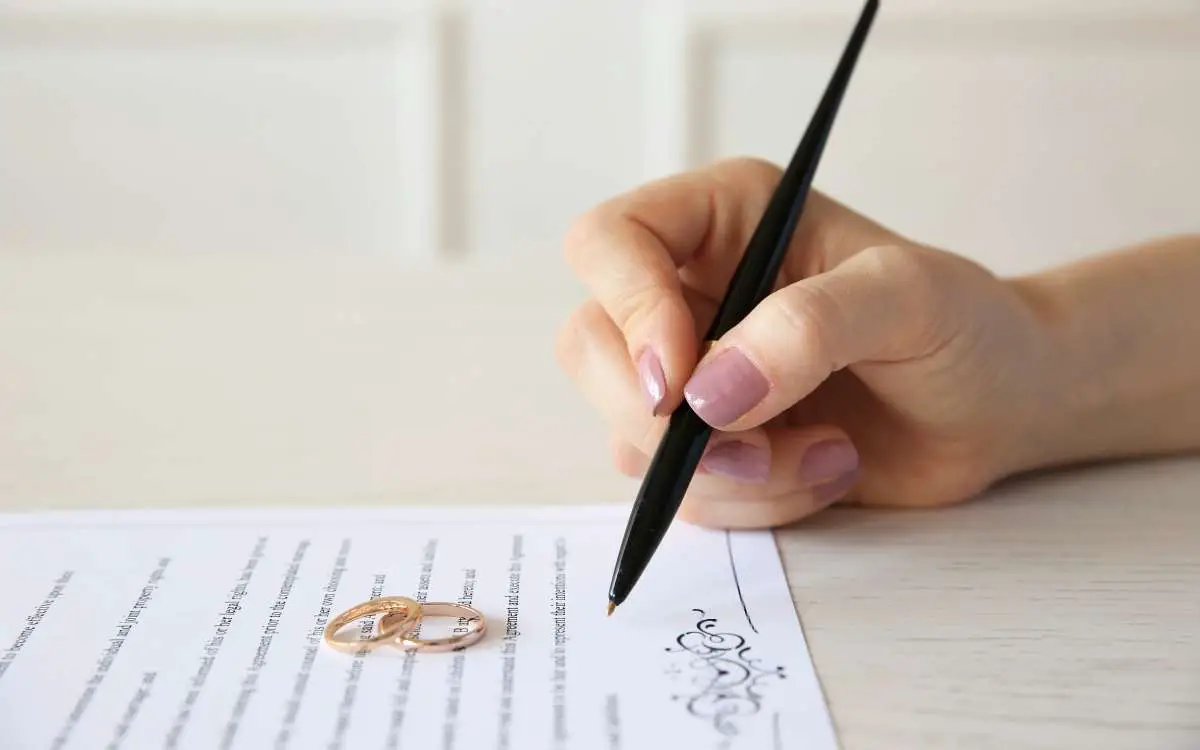 Photo of a hand holding a pen signing a document with a pair of rings on the paper