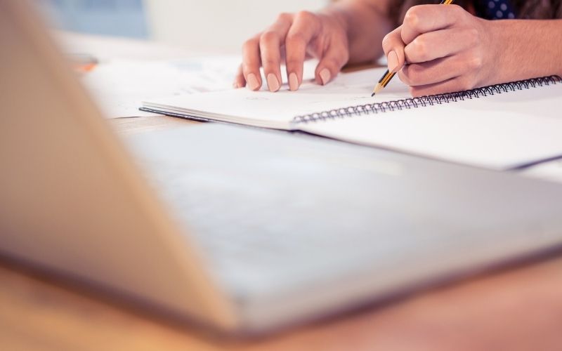 Photo of a pair of hands writing on a notebook in front of the laptop